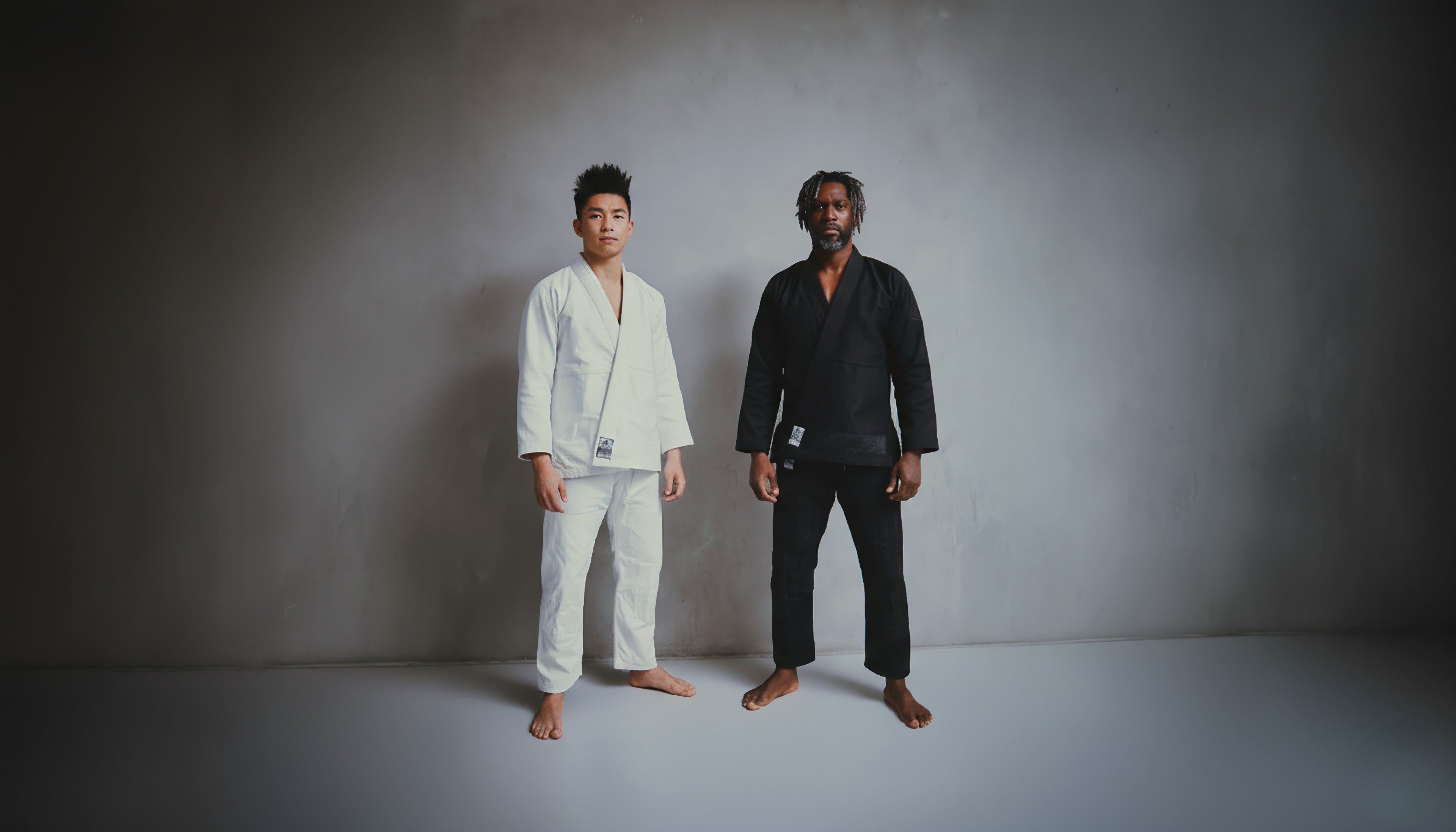 Two individuals, one in a white martial arts uniform and the other in black, standing against a plain background.